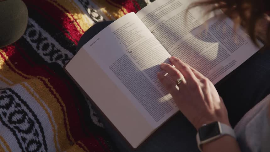 Woman with brown hair sits on a carpet and reads a book outdoors