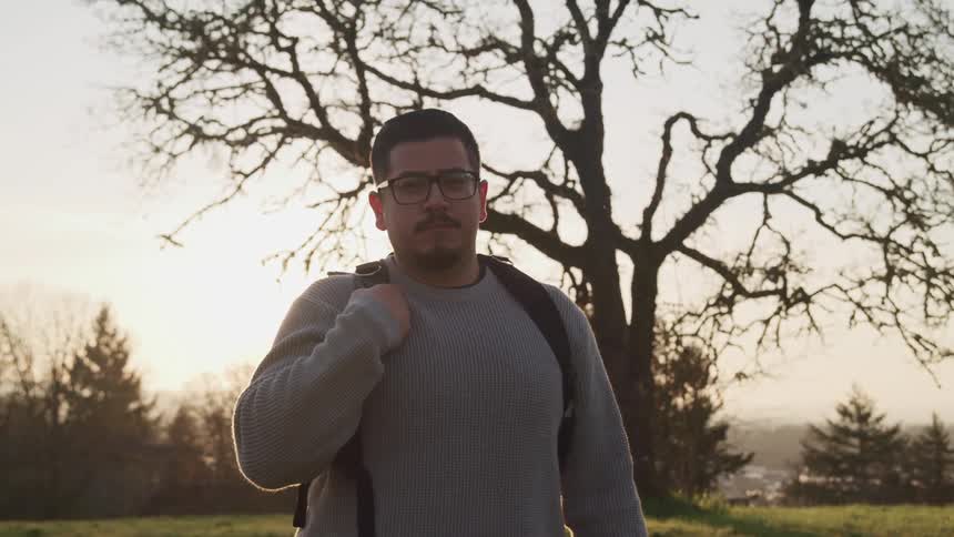 Chubby man with a long sleeve grey t shirt a backpack and glasses faces the camera with eye contact at sunset