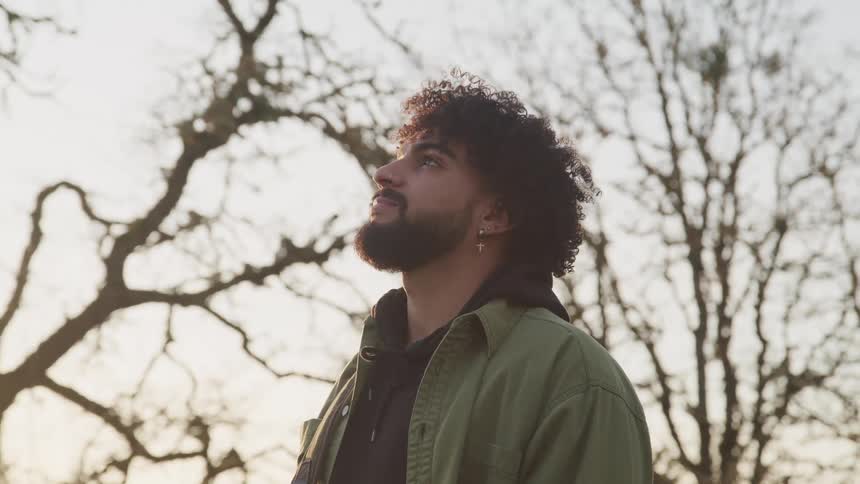 Side view of man with earrings curly hair and a beard looking up at the sky with bare trees in the background