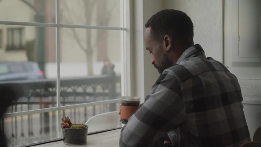 Black man with a beard and a checkered shirt drinks a glass of coffee in a cafe