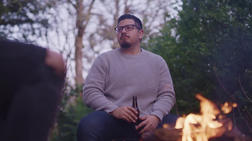 A man sits at a campfire holding a beer bottle