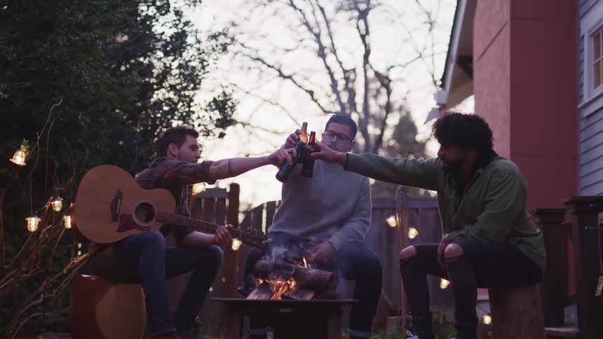 Friends hanging out around backyard fire pit cheering with beers