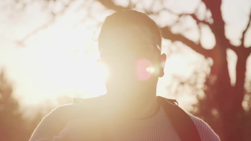 A man is standing outdoors at sunset while wearing a backpack