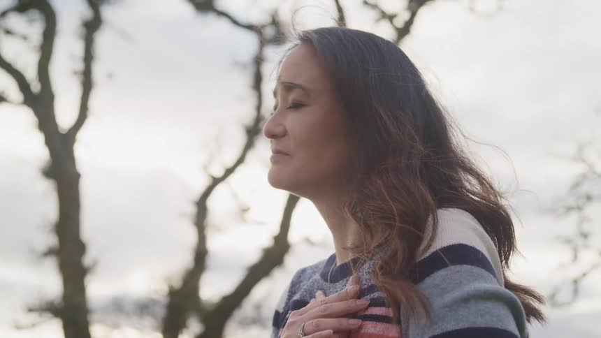 A woman stands outside with her eyes closed and her hands on her chest