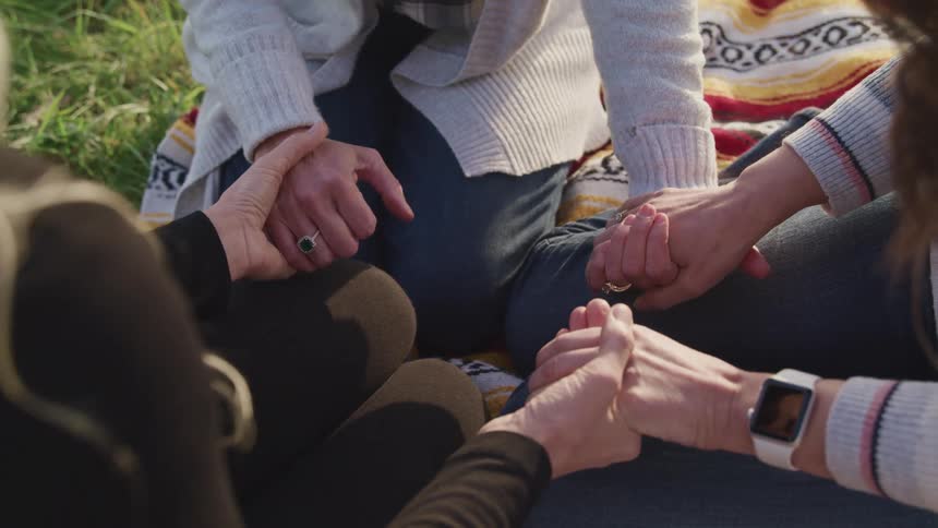 A group of three women hold hands while sitting in a circle on a blanket in a field during the day