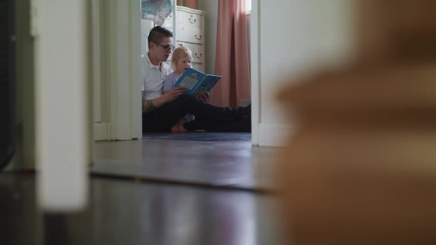 A man moves his arms while reading to his daughter as she sits on his lap on the floor of her bedroom