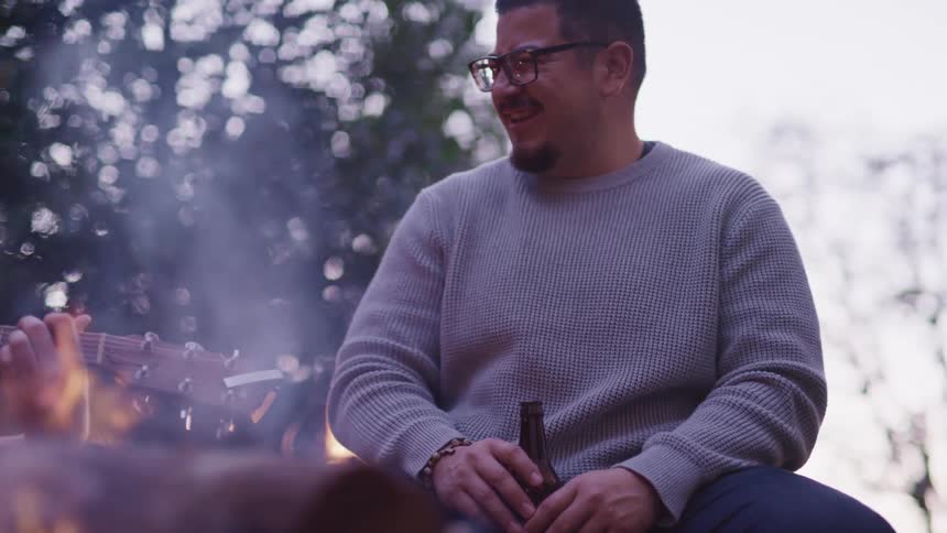 A man sitting and laughing near a bonfire with trees and the sky in the background