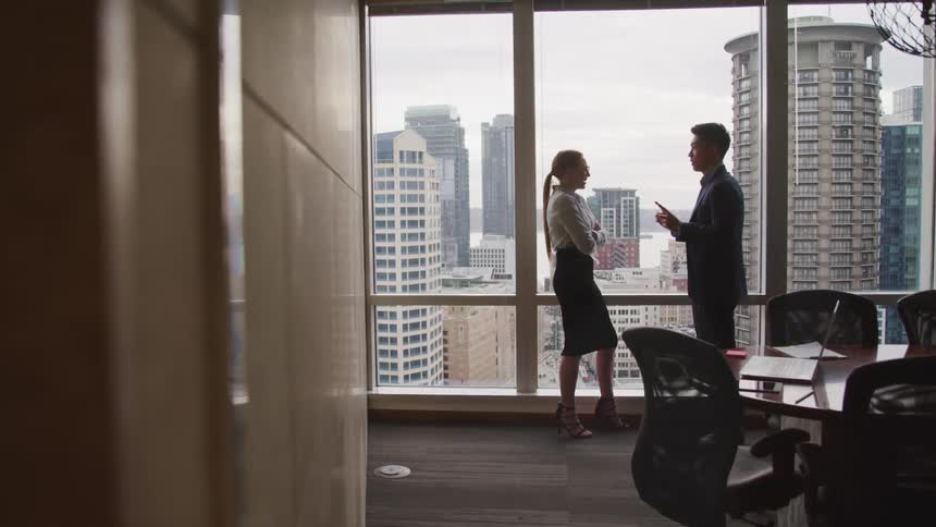 Business woman with a red ponytail a shirt and a black skirt speaks to asian businessman with a suit in office
