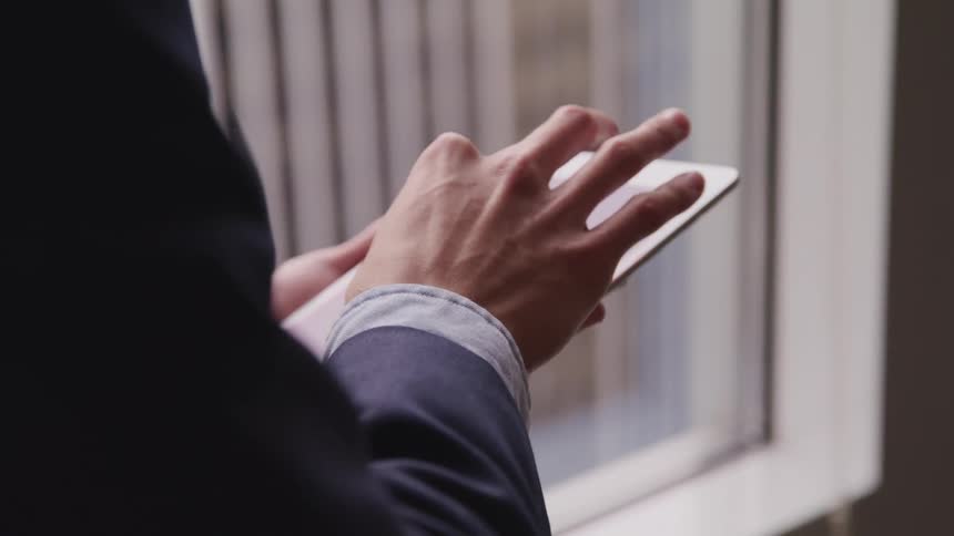 Business man in suit stands by office window and uses a tablet in the office