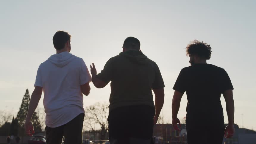 Three teenage boys walk across the grass towards a park as they toss a football and talk