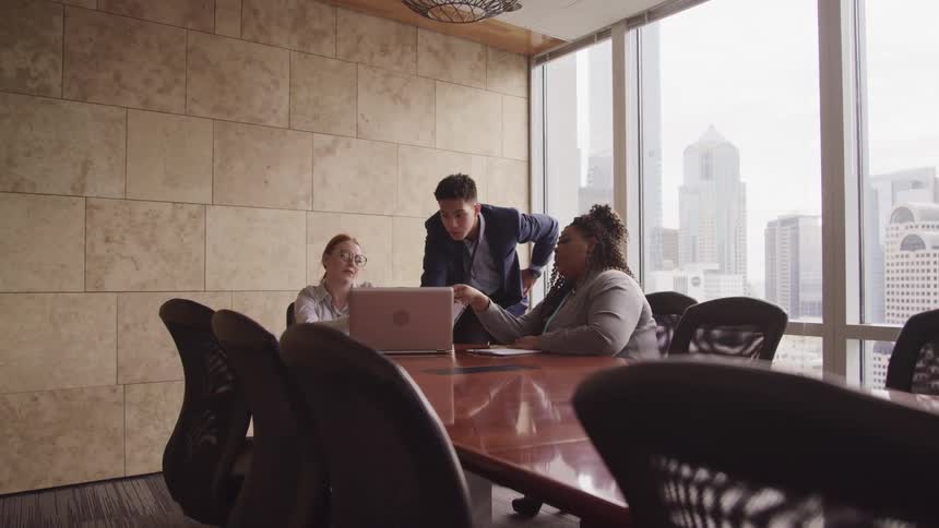 A man and two women around a conference table looking at a laptop with windows in the background