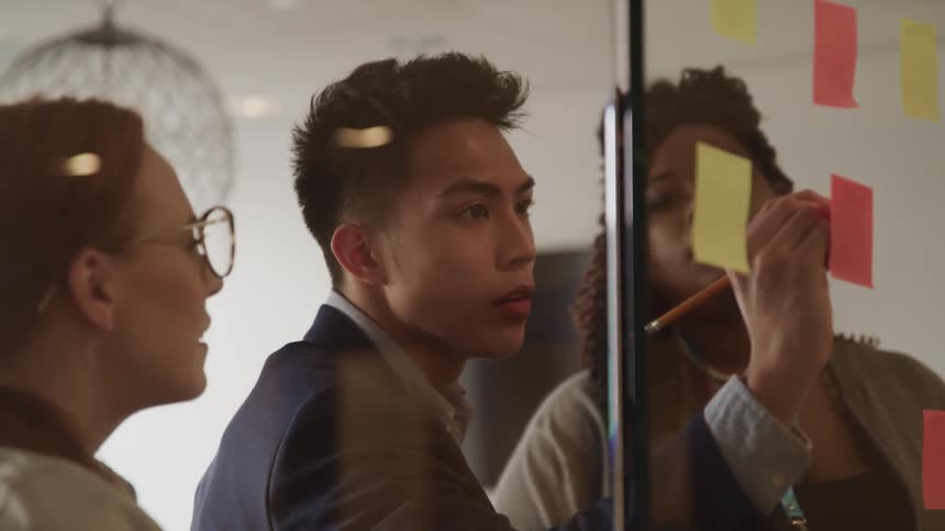 A man with a pencil writing on a sticky note on a glass as two women look on