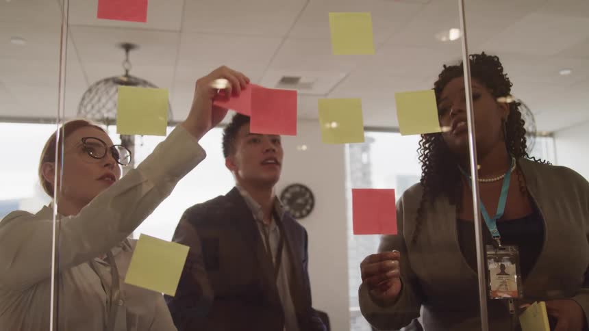 Three coworkers place sticky notes on a glass wall in an office