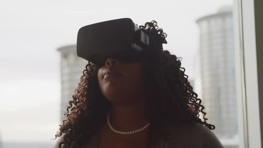 A black woman with virtual reality goggles on stands in a room with condos in the background
