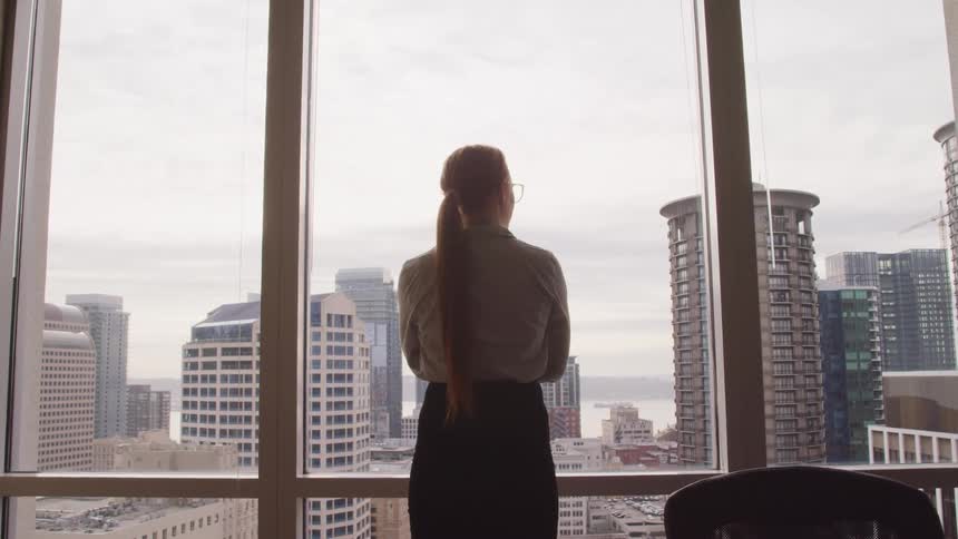 Woman stands at office window looking out at city skyscrapers