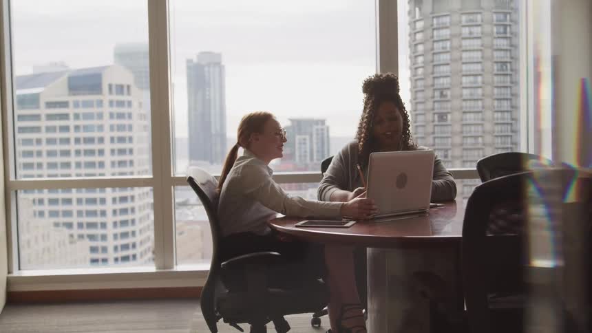 White businesswoman with ponytail glasses and a shirt and black woman sit at a table in the office with city skyline seen from the office windows
