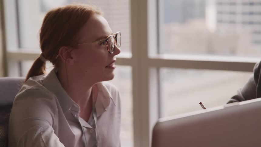 Woman with red hair glasses and a shirt sits at table and talks to co worker in the office