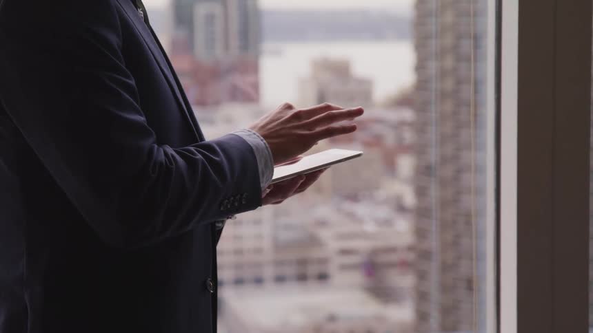 Business man in suit stands by office window and uses a tablet in the office