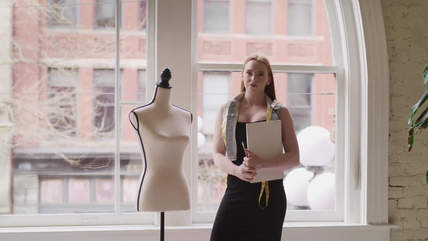 A woman standing next to a manikin holding a notebook and a pencil with a window in the background