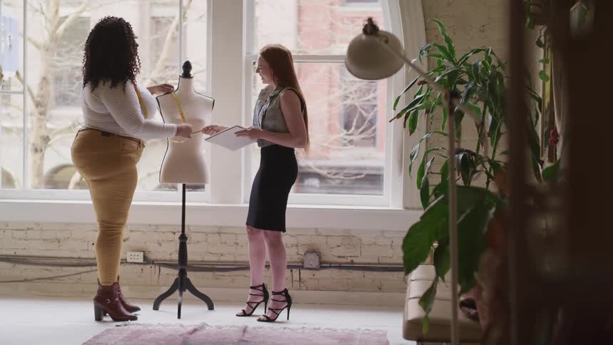 Two female fashion designers talk and laugh while measuring a mannequin in a studio