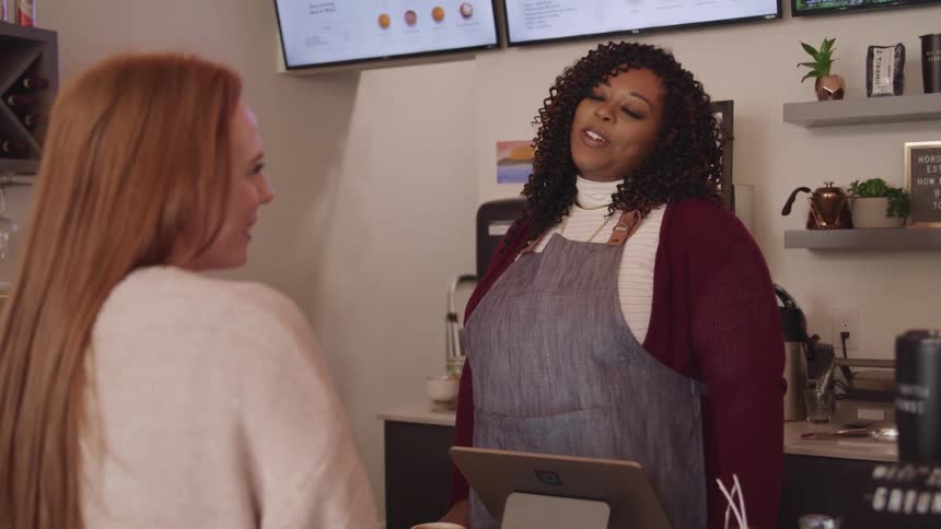 A woman standing with an apron behind the counter in a shop talking to another woman
