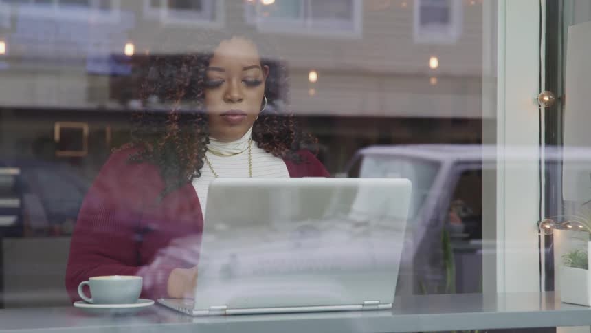 Woman sits in front of laptop near restaurant window with coffee cup at her side as passing traffic is reflected in glass