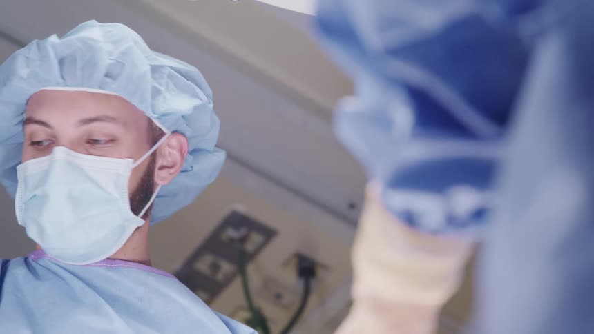 Man in surgical garb assists a second person who is sewing something