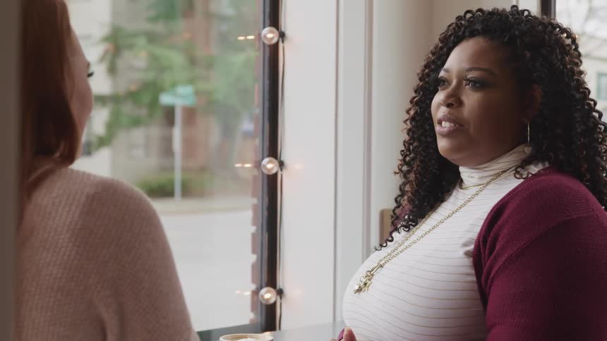 White woman with red hair a pink sweater and black woman with curly hair earrings sitting at a table drink coffee and talk to each other in a cafe