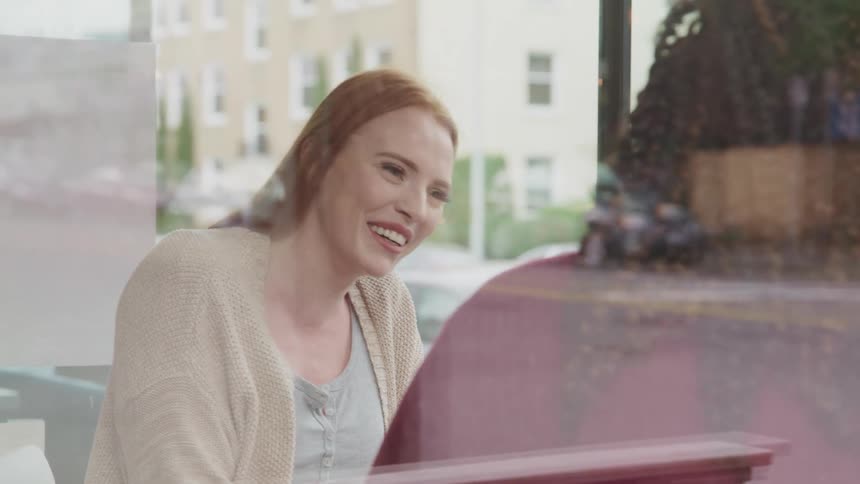 White woman with red hair a pink sweater and black woman with curly hair earrings sitting at a table drink coffee and talk to each other in a cafe