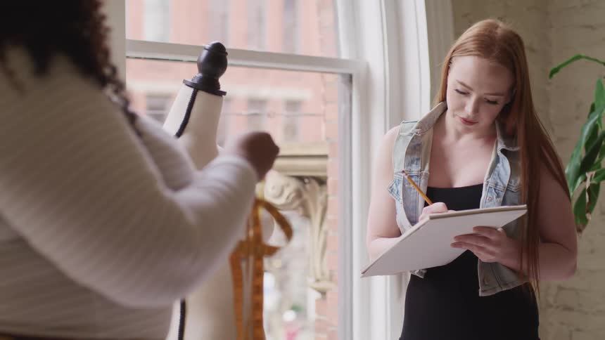 A woman stands near a mannequin in a shop while writing in a notebook as another standing near her unravels a measuring tape