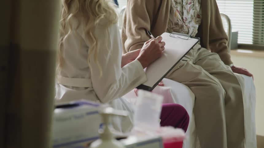 A female doctor writes on a clipboard while sitting on a chair next to an elderly patient sitting on a hospital bed