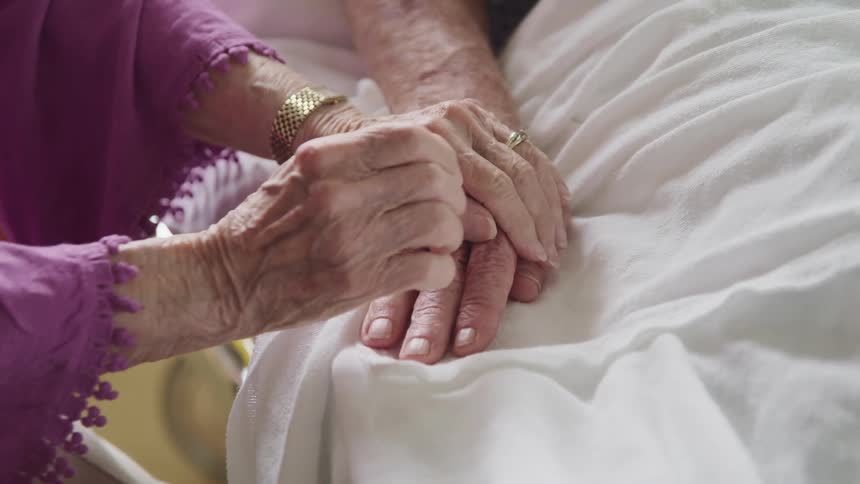 A woman holding a mans hand laying on a bed with sheets