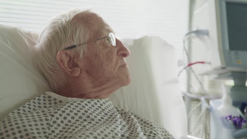 An elderly man sitting in a hospital bed looks around the room