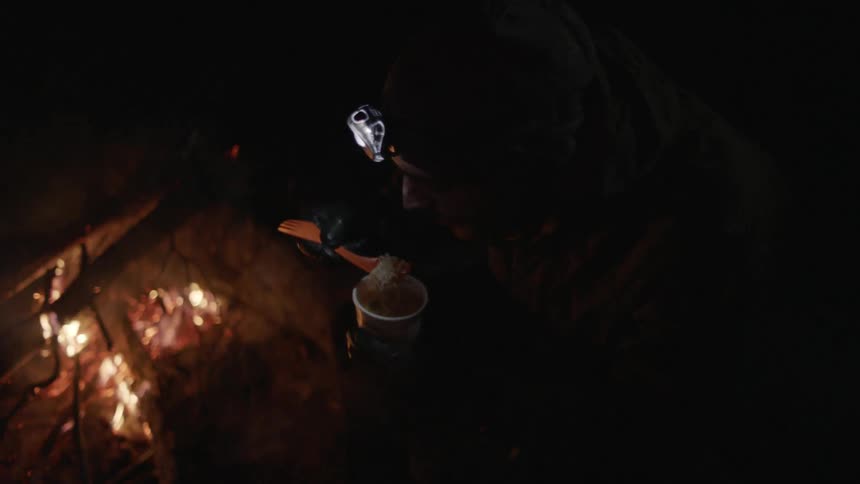A man is eating from a cup by a fire