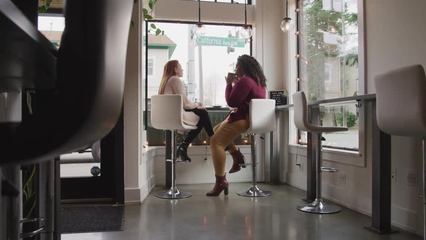 A woman drinks from a coffee mug as she sits with another woman and they chat in a cafe during the day