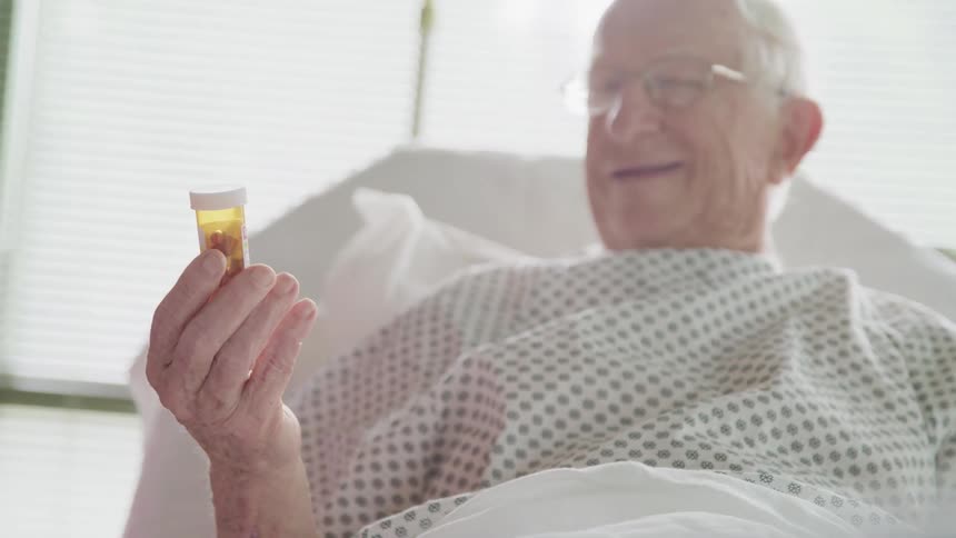 Elderly man smiles and holds a pill bottle while laying in a hospital bed