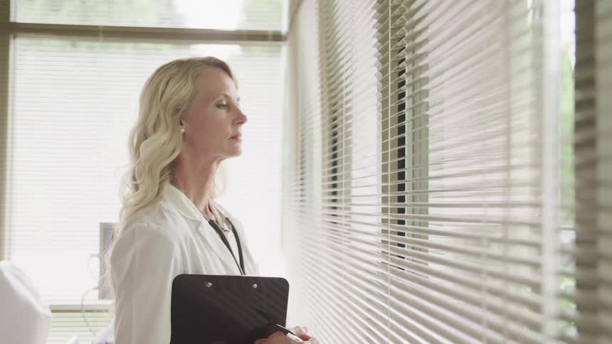 A woman wearing a white jacket stares out windows of a hospital room