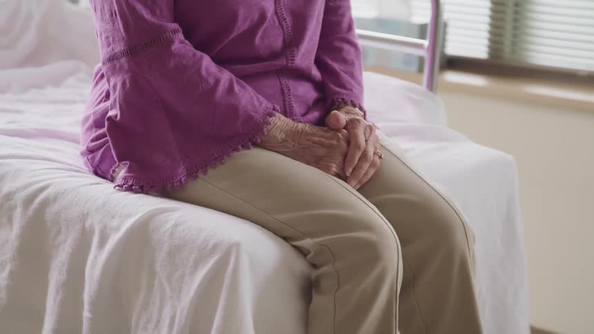 Elderly woman sits patiently with her hands over her legs on a bed in a hospital