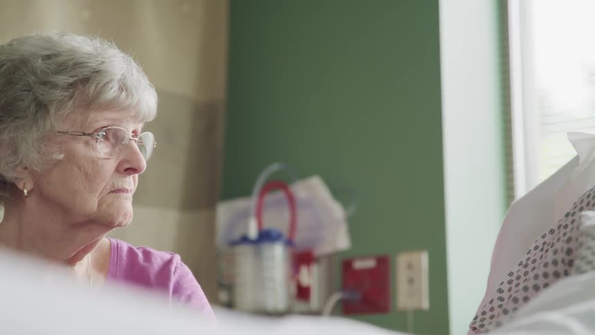 An elderly woman sits beside a patient in a hospital bed looking sad