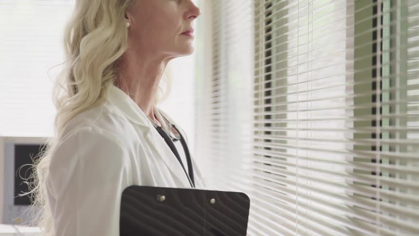 Side view of a female doctor with blonde hair and a stethoscope around her neck looking out the window in a clinic