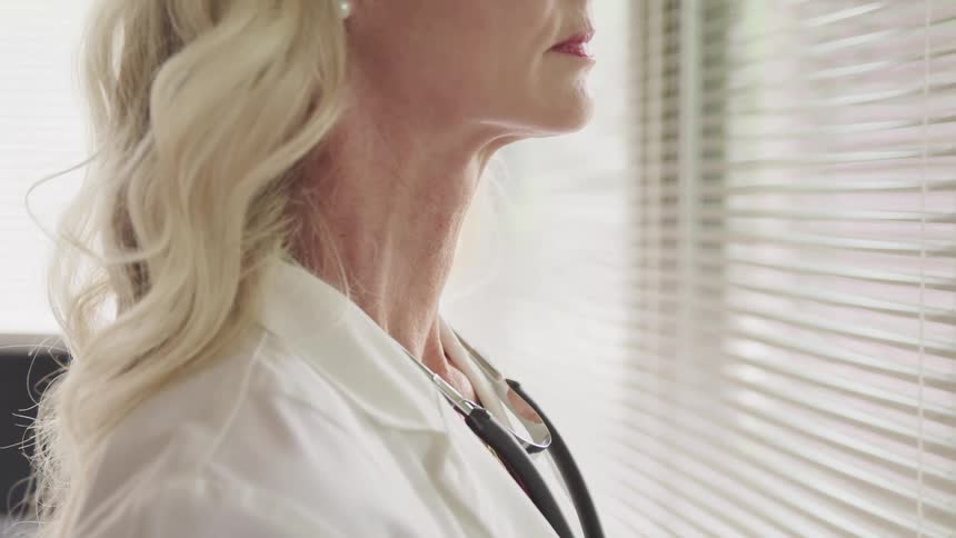 A woman with a white shirt and a stethoscope standing in front of a window with blinds