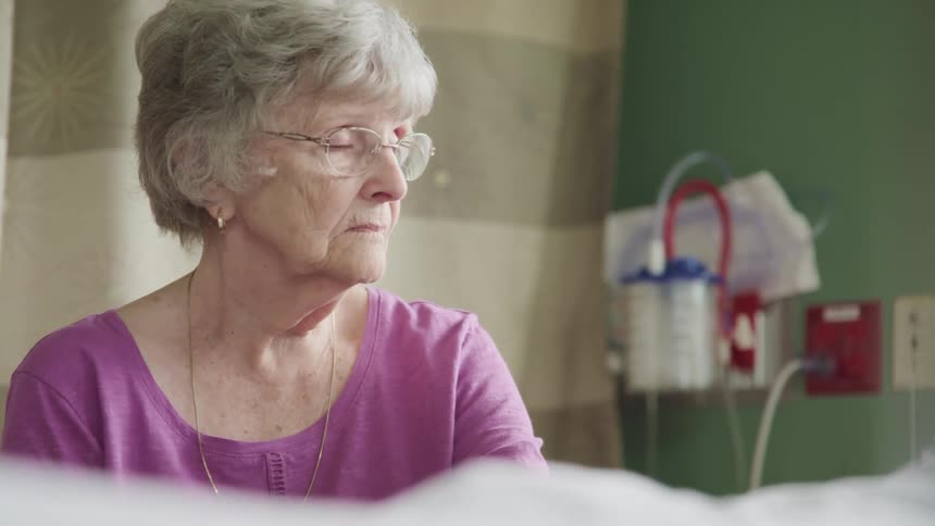 Elderly woman sits by patients bed in hospital room
