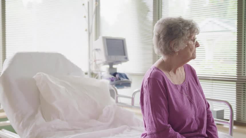 An elderly woman sits on a hospital bed and gazes out of the window
