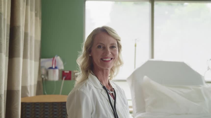 Blonde middle aged female doctor with a doctor lab and a stethoscope sits next to a stretcher and beams at the camera in the clinic