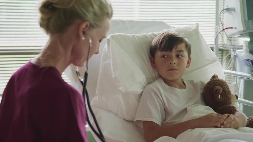 A nurse sitting beside a hospital bed containing a boy adjusts her stethoscope in her ear