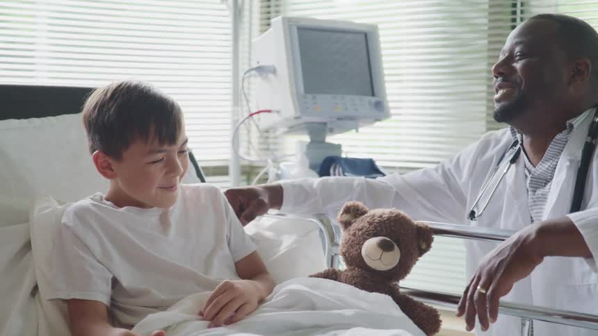 A doctor kneels beside a hospital bed and jokes with the young patient resting there