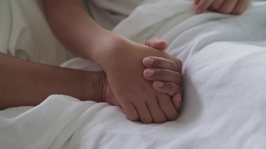 Person holds the hand of another lying in bed with sheet over them