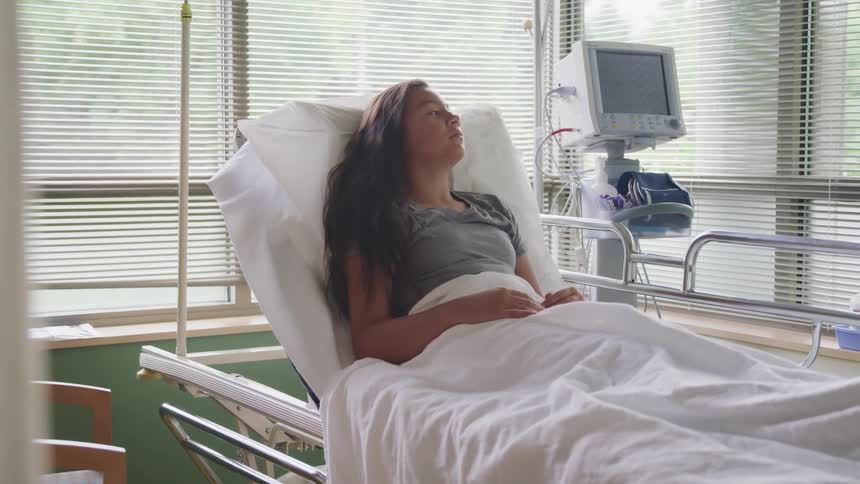 A teenage girl lays in a hospital bed looking out the window