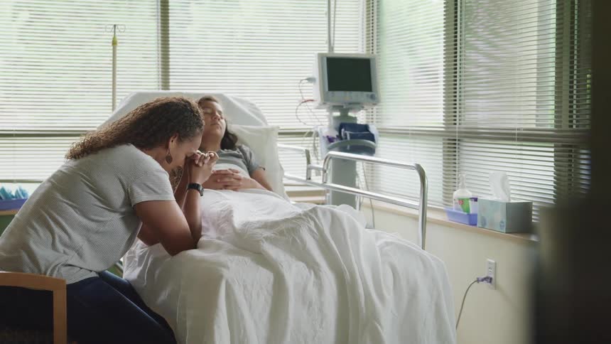 A woman leans against the hospital bed that her teenager daughter is laying in with her head leaning on her hands as if in prayer