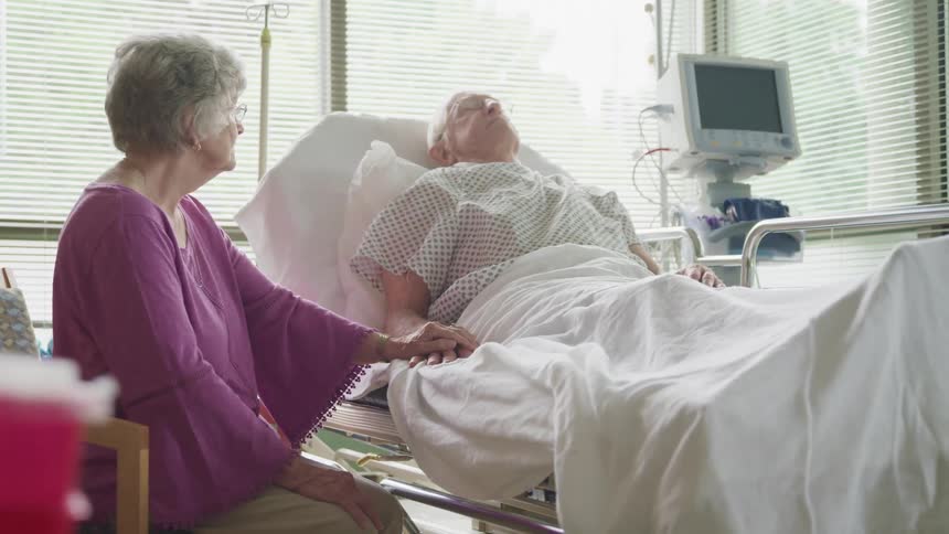 A man lies in a hospital bed and sleeps as his wife sits next to him and holds his hand